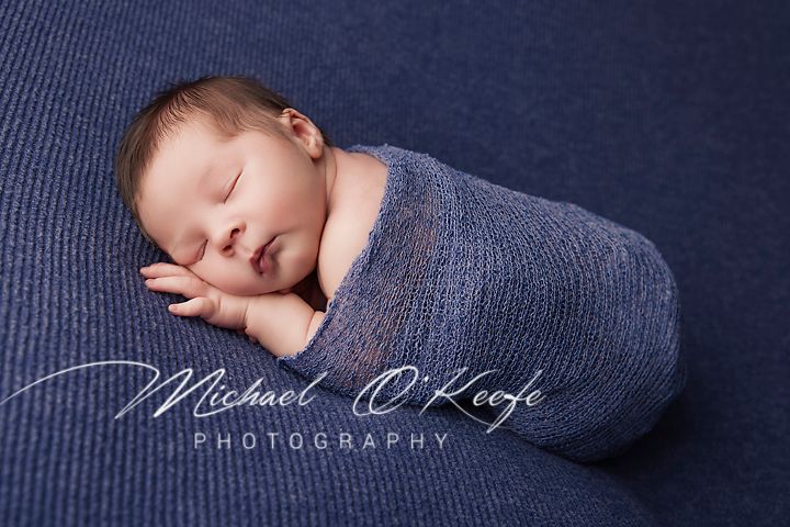 Sleeping newborn baby wrapped in soft blue fabric against navy textured background in professional portrait series.