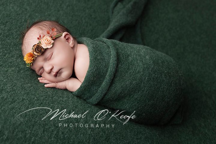 Newborn baby swaddled in green wrap with floral headband, sleeping peacefully in professional photography portrait.