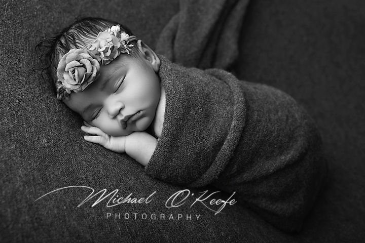 Black and white newborn photo of sleeping baby wrapped in gray swaddle wearing floral headband by Michael O'Keeffe Photography.