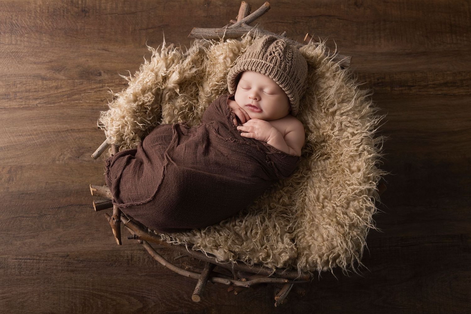 Newborn baby sleeping peacefully in a rustic basket wrapped in brown cloth wearing a knit bear hat.