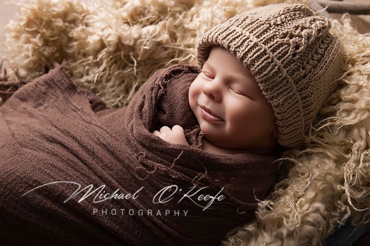 Sleeping newborn baby wrapped in brown knit blanket and hat, posed on fluffy cream rug, by Michael OKeefe Photography.