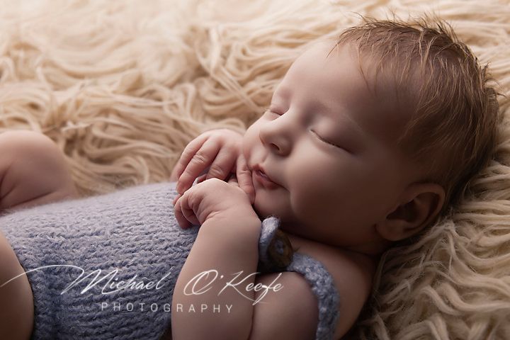 Sleeping newborn baby in lavender knit outfit on fluffy cream fur, photographed by Michael O'Keefe Photography.
