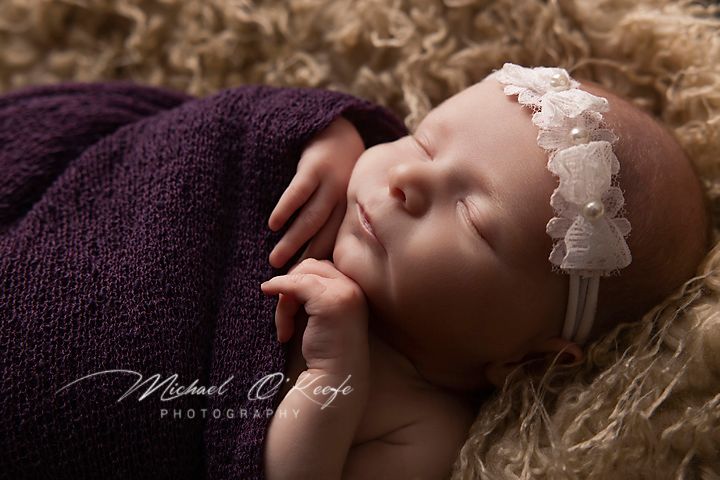 Sleeping newborn wearing purple wrap and white lace headband rests on soft brown fur backdrop during professional photoshoot.