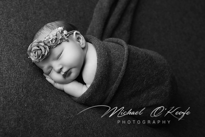 Black and white newborn photo of sleeping baby wrapped in gray swaddle wearing floral headband by Michael O'Keefe Photography.
