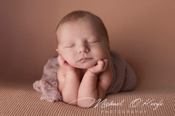 Sweet newborn baby portrait with hands under chin in soft pink lighting.