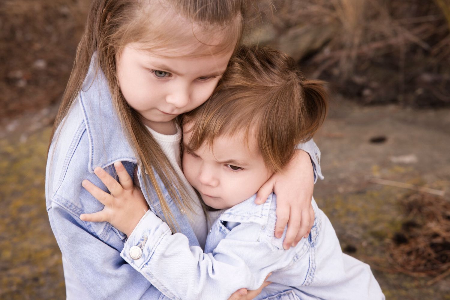 Two children in light colored clothing sharing a tender embrace outdoors.