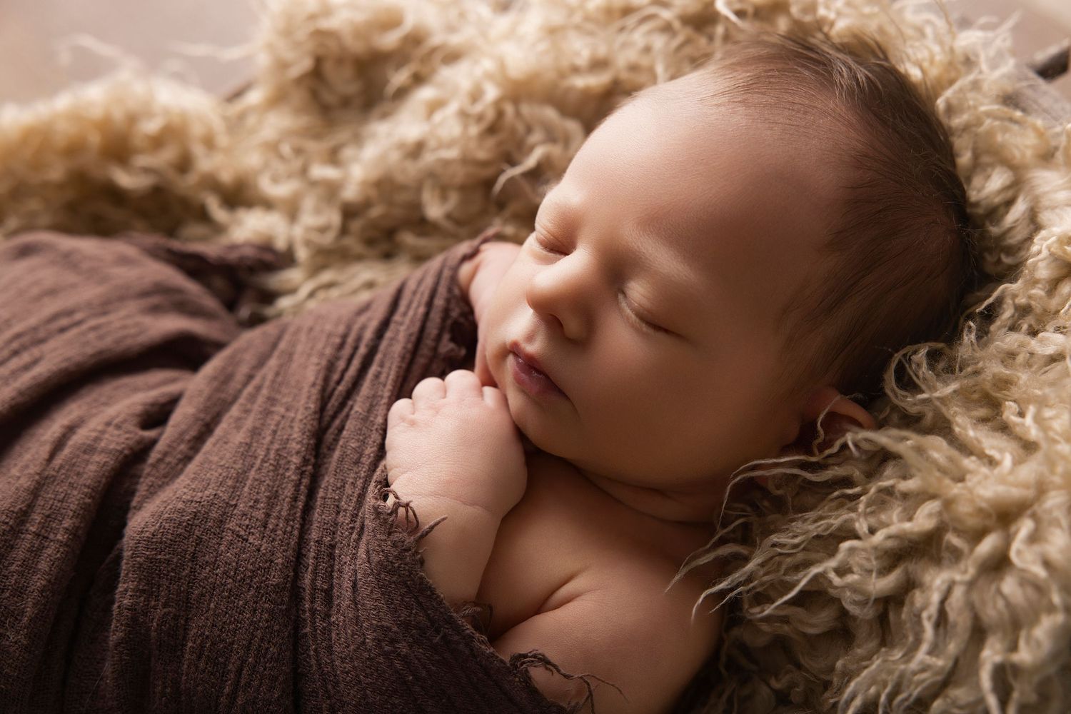 Peaceful newborn baby swaddled in brown wrap sleeping on fluffy tan fur blanket in professional portrait.