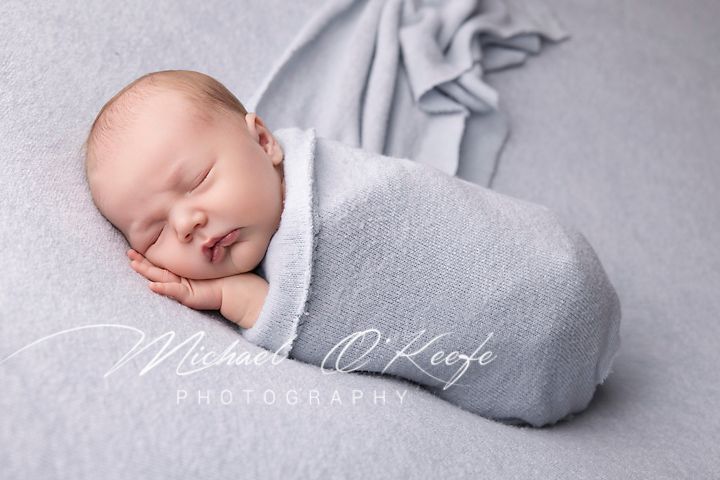 A sleeping newborn baby wrapped snugly in a soft gray blanket lies peacefully on a matching gray backdrop.