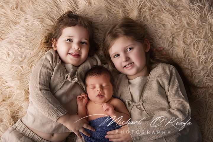Two smiling young girls cuddle a sleeping newborn baby on a soft cream fur blanket in a studio portrait.