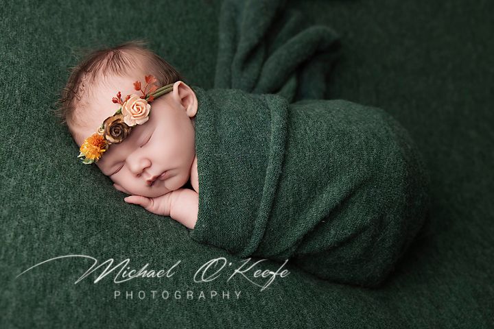 A sleeping newborn tenderly swaddled in soft dark green fabric with a delicate floral headpiece.