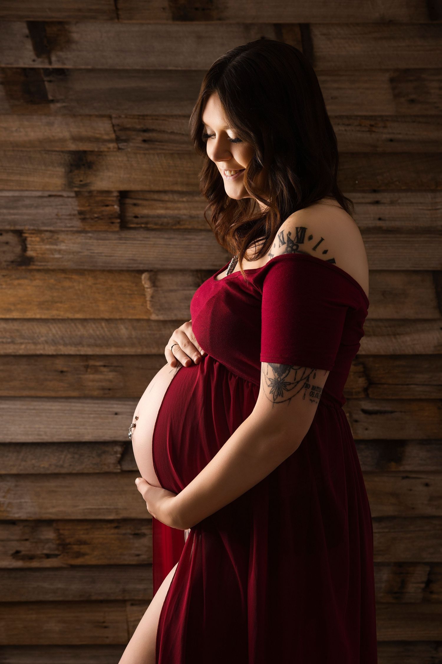 A pregnant woman in a burgundy velvet dress poses against a rustic wooden wall in a maternity photoshoot series.