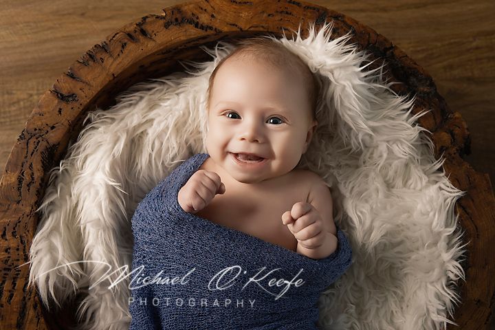 Smiling baby wrapped in blue, nestled in a fluffy white fur bowl, captured by Michael O'Keefe Photography.