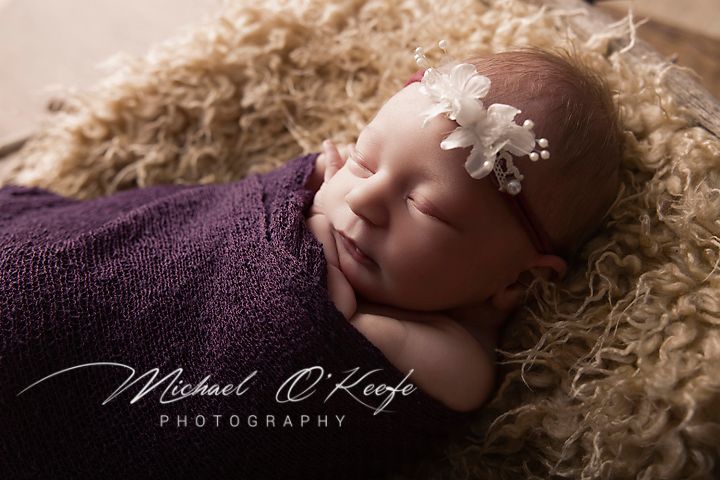 Newborn baby swaddled in purple wrap with white floral headband, sleeping on fluffy tan fur by Michael O'Keefe Photography.