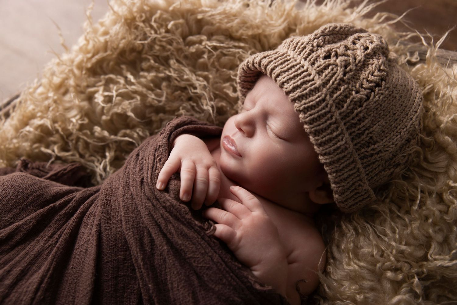 Sleeping newborn wrapped in brown fabric wearing knit beanie on textured backdrop.