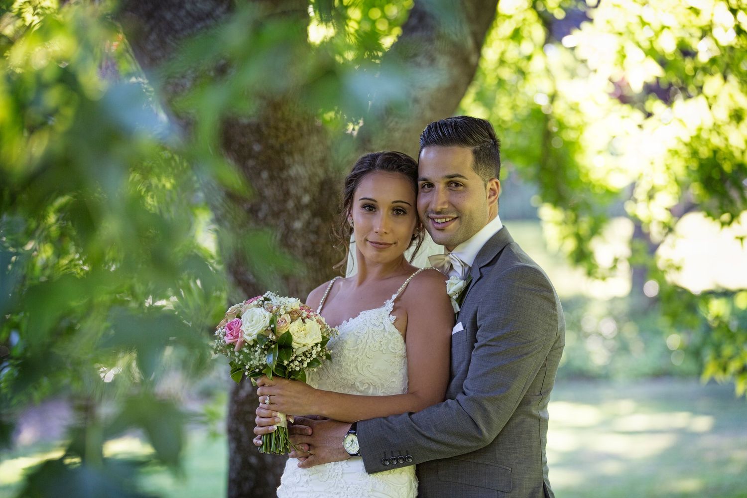 A romantic wedding portrait taken in a garden setting with natural sunlight filtering through trees.