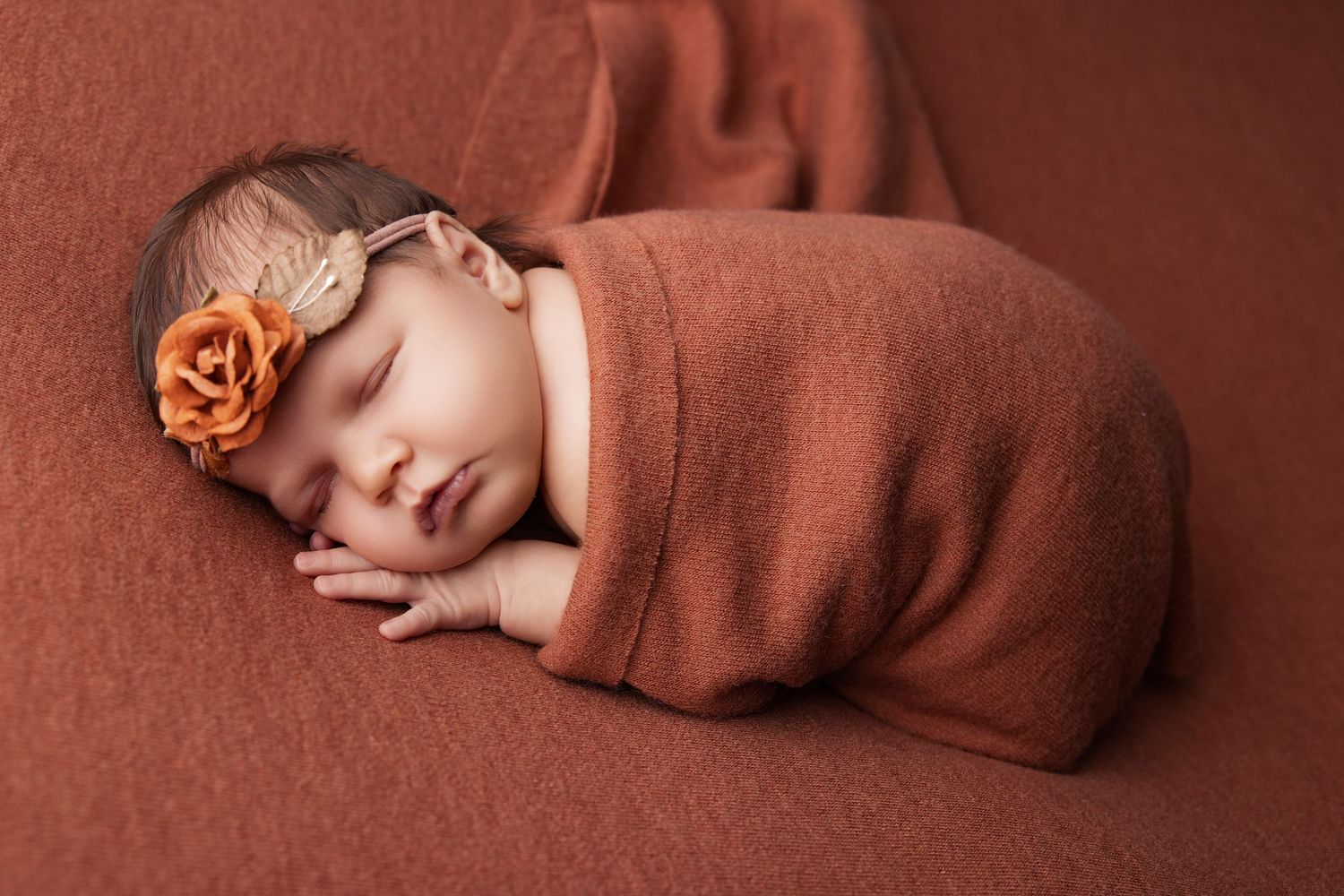 Sleeping newborn wrapped in rust-colored blanket with matching flower headband.