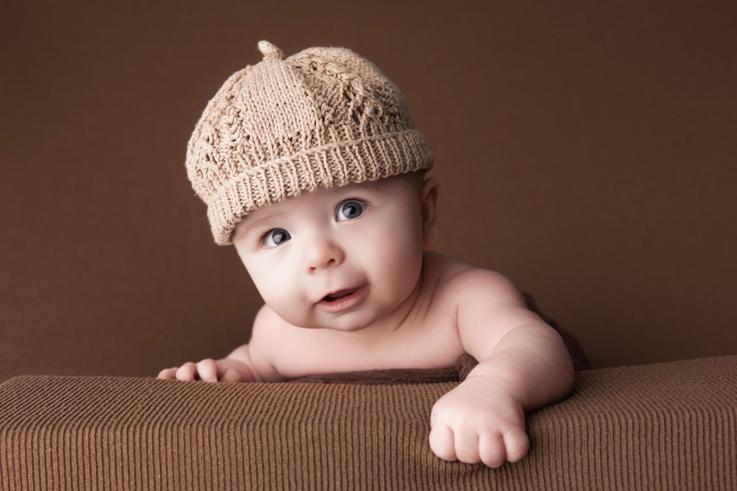 Baby wearing beige knit cap lying on brown textured surface.