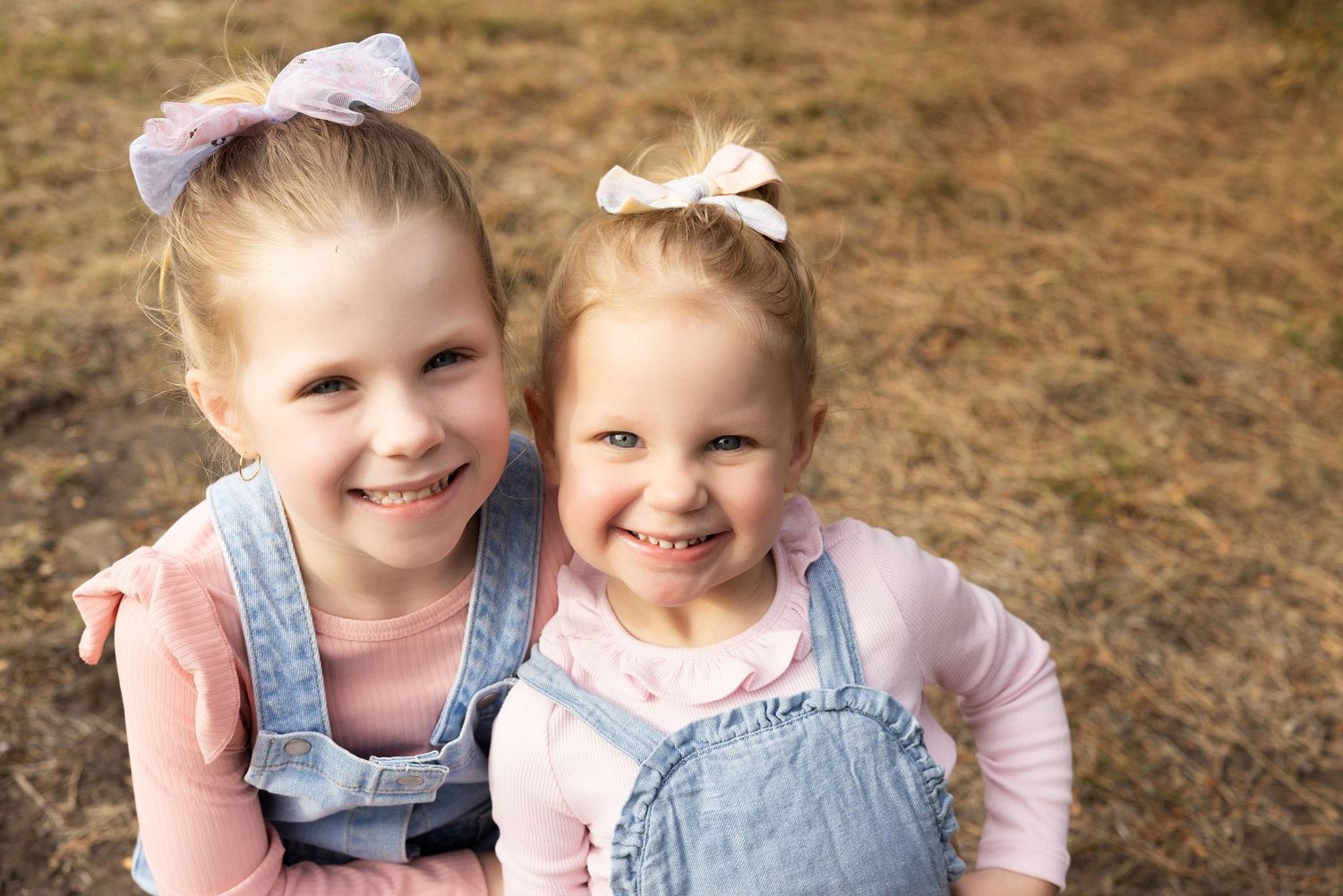 Two young sisters in matching hair bows and denim outfits smile brightly outdoors.