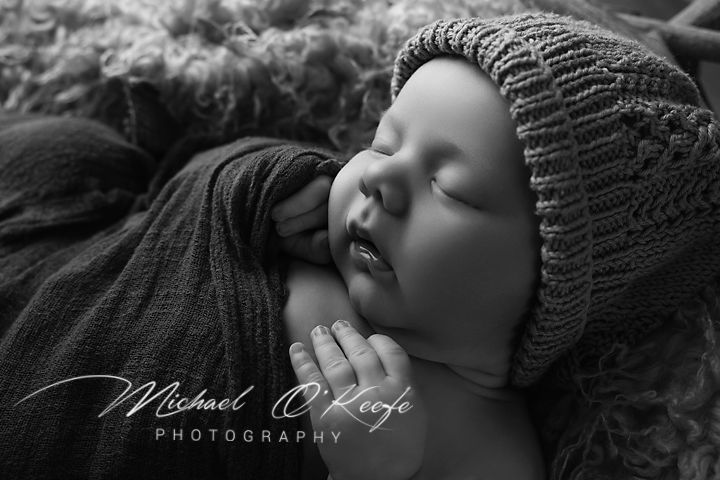 Black and white newborn photo of sleeping baby in knit bonnet, wrapped in soft fabric by Michael O'Keefe Photography.