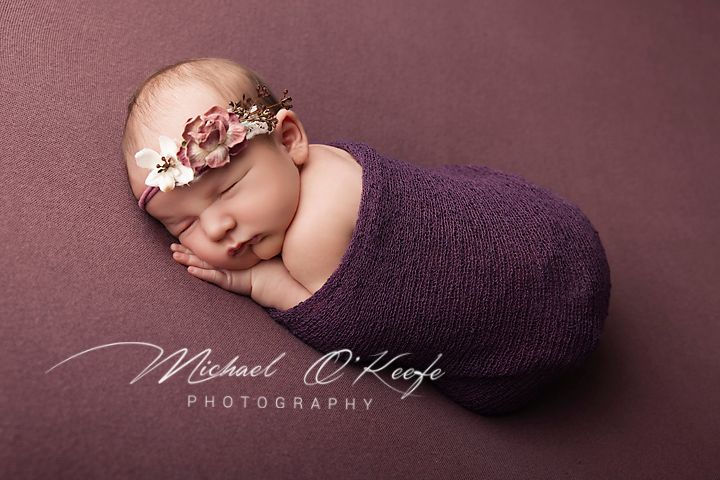 Newborn baby swaddled in purple wrap with floral headband, sleeping peacefully on mauve background by Michael O'Keefe Photography.