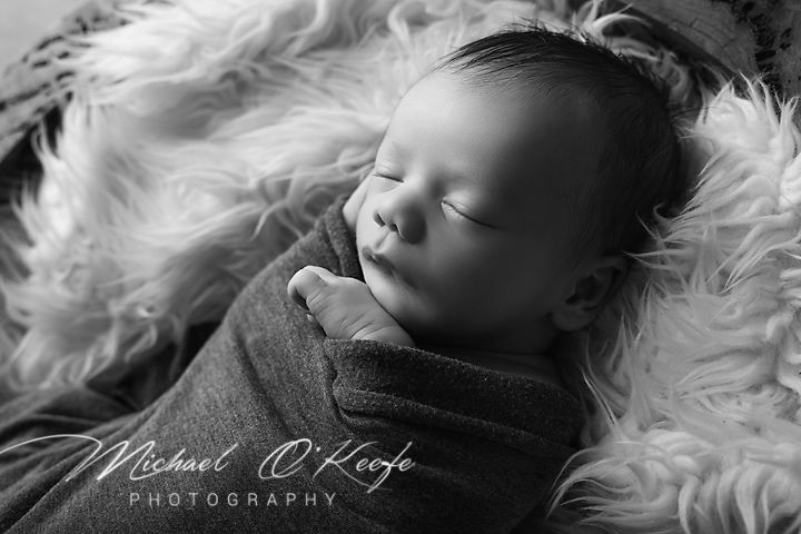 Black and white newborn photo of a sleeping baby wrapped in a soft blanket on a fluffy rug by Michael O'Keefe Photography.