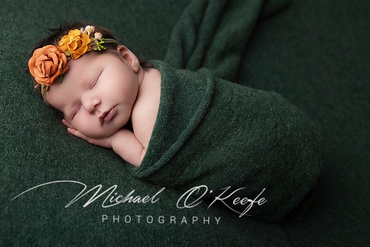 Newborn baby wrapped in green knit swaddle with orange floral headband, sleeping peacefully in professional photography portrait.