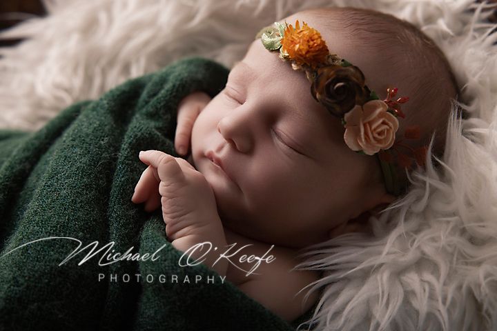 Sleeping newborn baby wearing a floral headband, wrapped in green fabric on a fluffy white blanket.