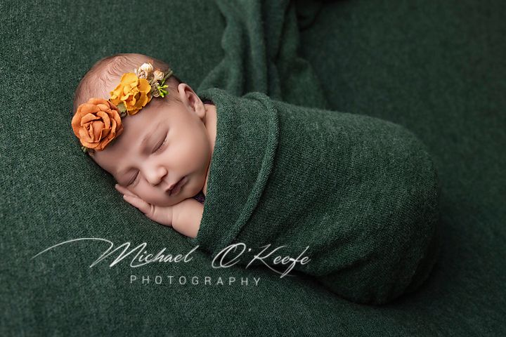 Newborn baby swaddled in green wrap with orange floral headband, sleeping peacefully in professional photo.