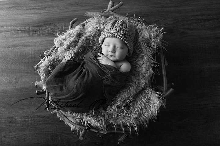 Black and white photo of a sleeping newborn baby in a knit hat nestled in a cozy basket with fur lining.