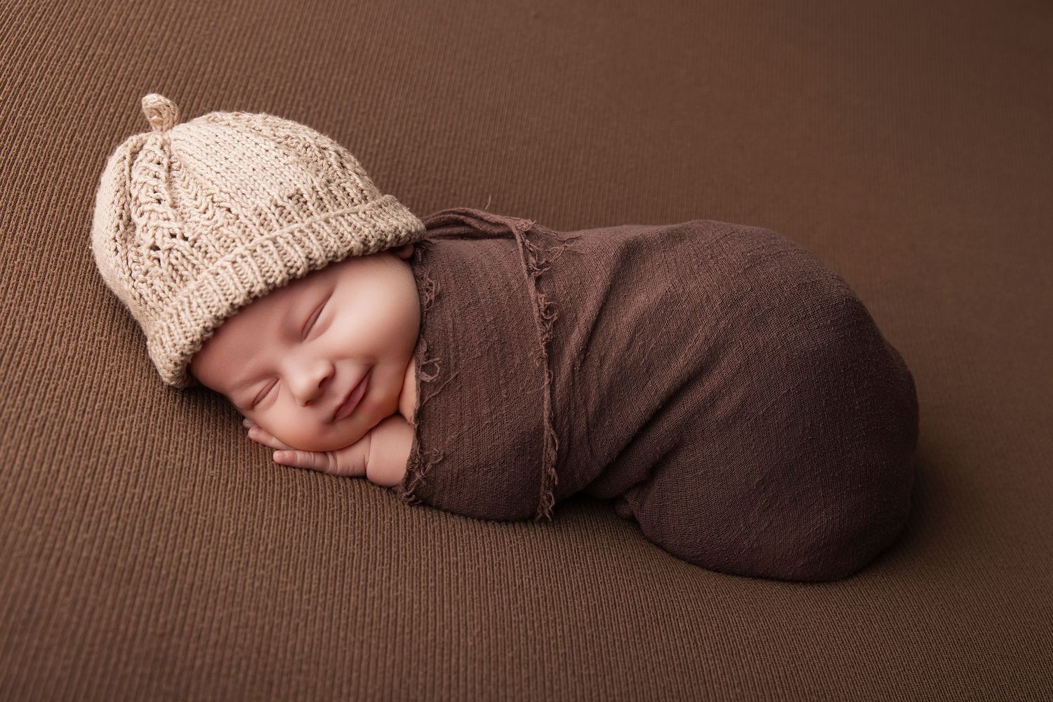 Smiling newborn baby swaddled in brown wrap wearing a knit beige hat, peacefully sleeping on brown background.