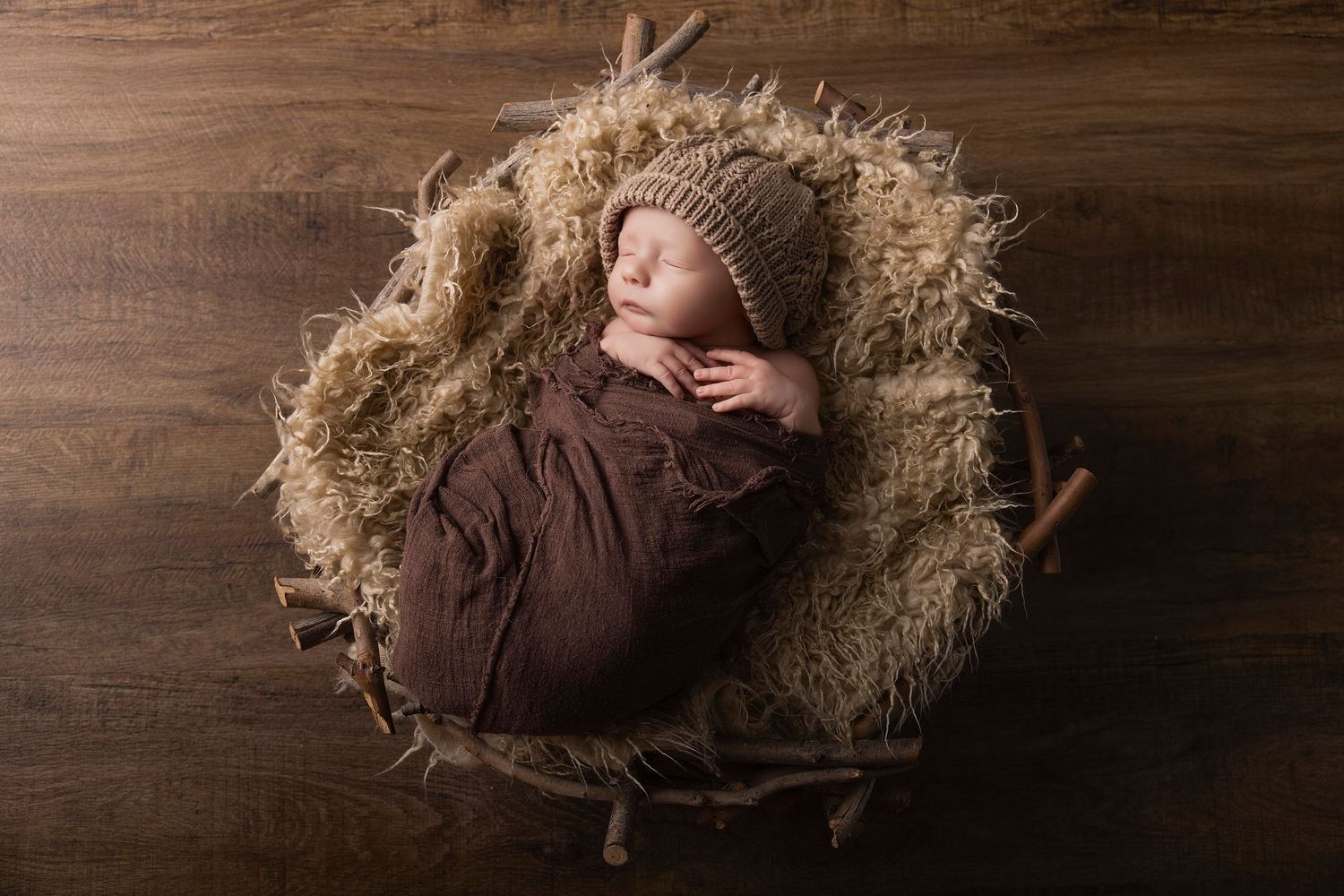 Newborn baby sleeping peacefully in a rustic basket with fur lining, wearing a brown knit hat, wrapped in brown fabric.