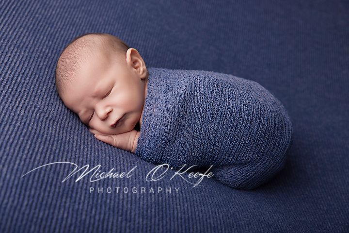 Sleeping newborn wrapped in soft blue swaddle blanket on textured navy background during professional photoshoot.