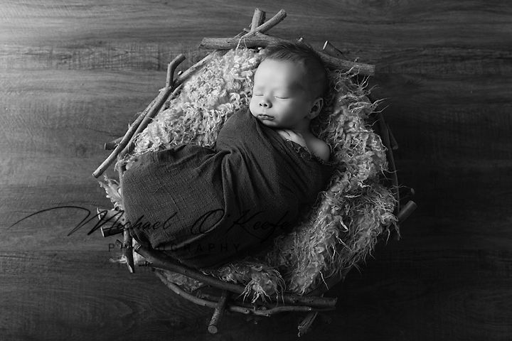 A sleeping newborn baby wrapped in black fabric lies in a rustic nest-like setting against a dark background.