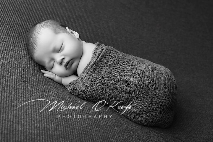 Black and white newborn photography of a sleeping baby wrapped snugly in a gray swaddle on textured background.