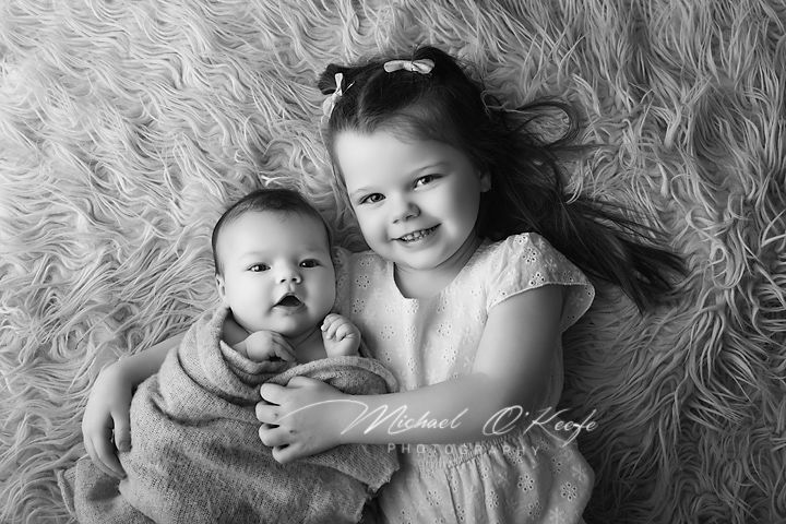 Black and white photo of a smiling toddler girl holding a newborn baby on a fluffy rug.