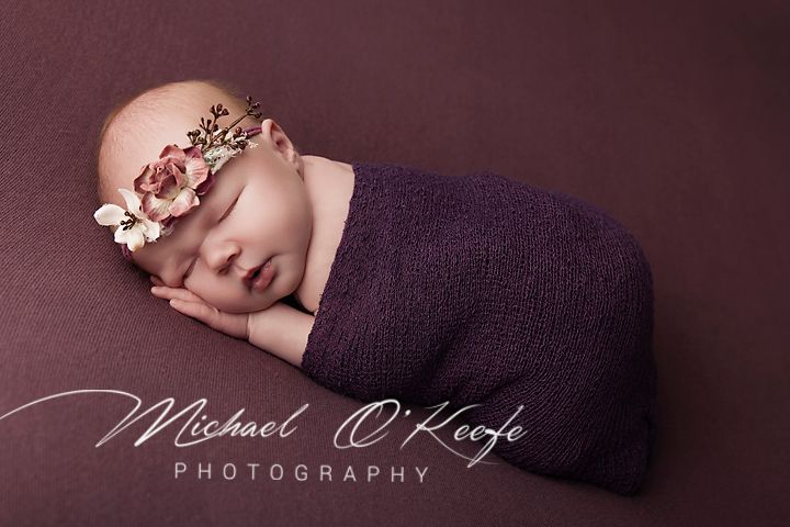 Newborn baby girl swaddled in purple wrap with floral headband, sleeping peacefully on mauve background.