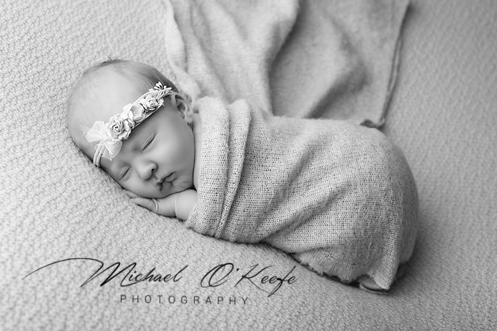 Black and white newborn photo of a sleeping baby wrapped in a soft blanket wearing a floral headband.