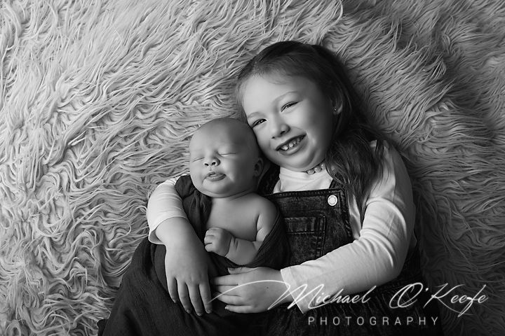 Black and white portrait of siblings lying on a fluffy rug, one wearing overalls and both smiling at the camera.