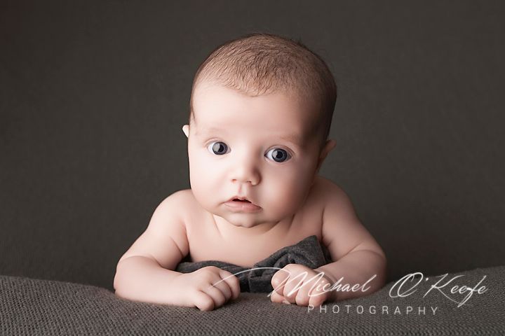 A baby lies on their tummy on a gray blanket, looking up with wide blue eyes in a professional studio portrait.