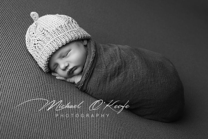 Black and white newborn photography of a sleeping baby wrapped in dark fabric wearing a white knitted beanie.