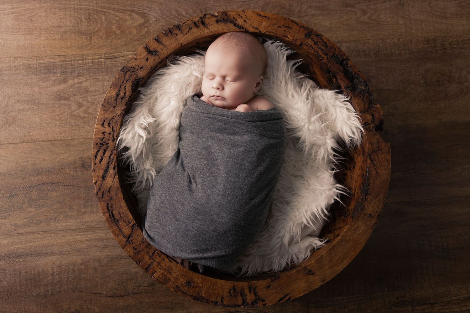 Sleeping newborn cradled in rustic wooden bowl with soft white padding.