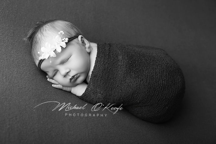 Black and white newborn photo of a sleeping baby swaddled in dark wrap with floral headband by Michael O'Keefe Photography.
