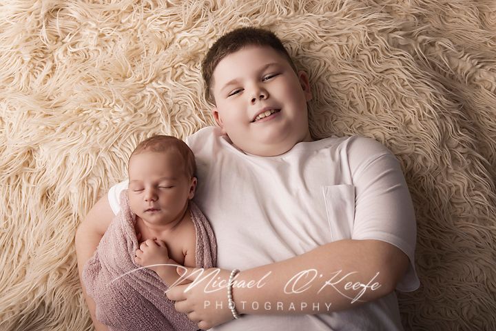 A smiling boy lies on a fluffy cream rug beside a sleeping newborn baby wrapped in a pink swaddle.