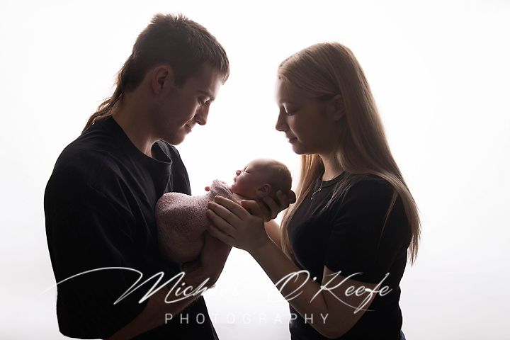 Silhouette of new parents cradling their newborn baby against a bright white background in a studio portrait.