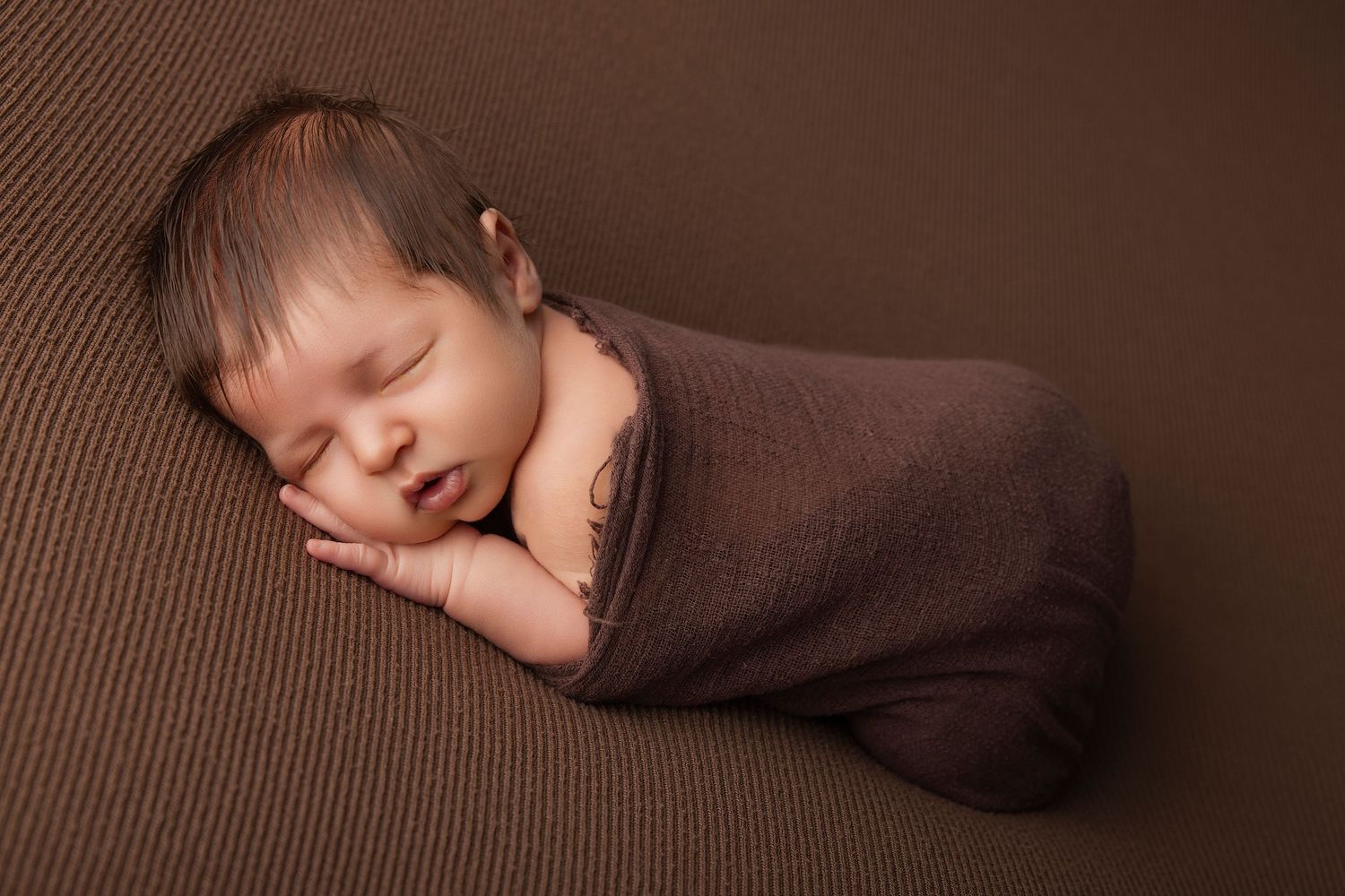 A peaceful newborn baby swaddled in a brown wrap, sleeping on a matching brown textured background.