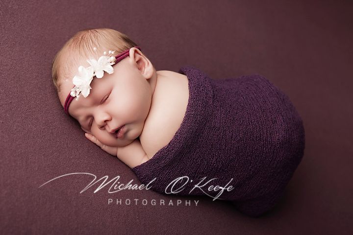 Newborn baby wrapped in purple knit swaddle with floral headband, sleeping peacefully on purple backdrop.