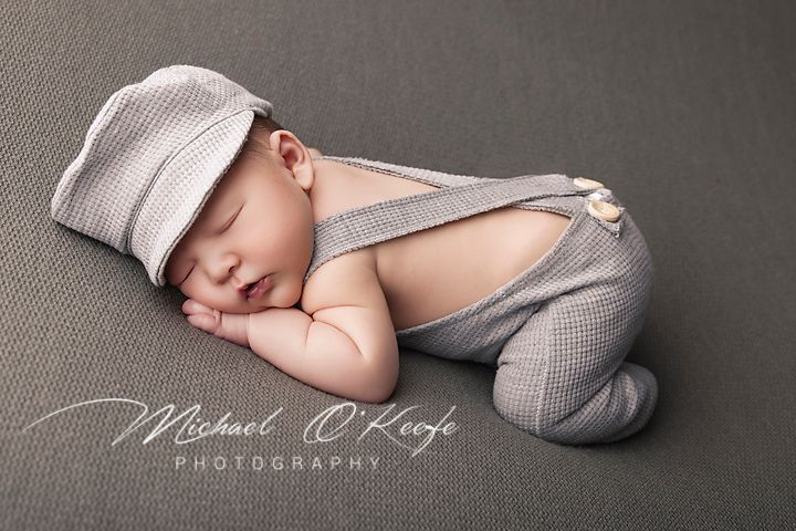 Sleeping newborn in gray overalls and newsboy cap curled up on soft gray backdrop during professional photo session.