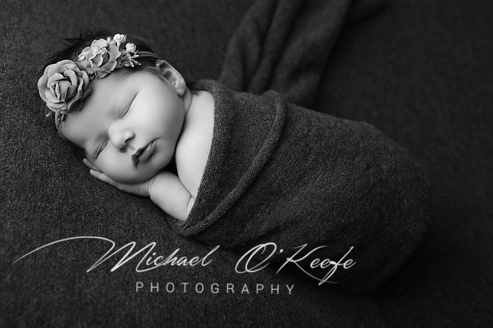 Black and white newborn portrait of a sleeping baby wrapped in a soft blanket with a floral headband by Michael O'Keefe Photography.