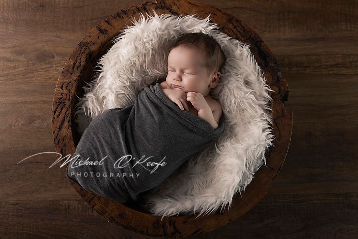 Newborn baby wrapped in gray swaddle sleeping peacefully in rustic wooden bowl with white fur, professional photography.