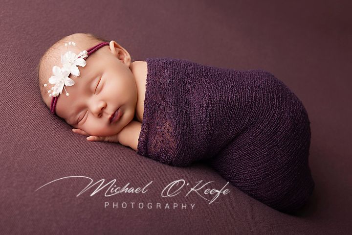 Newborn baby girl swaddled in purple wrap with white flower headband, sleeping peacefully on purple backdrop.